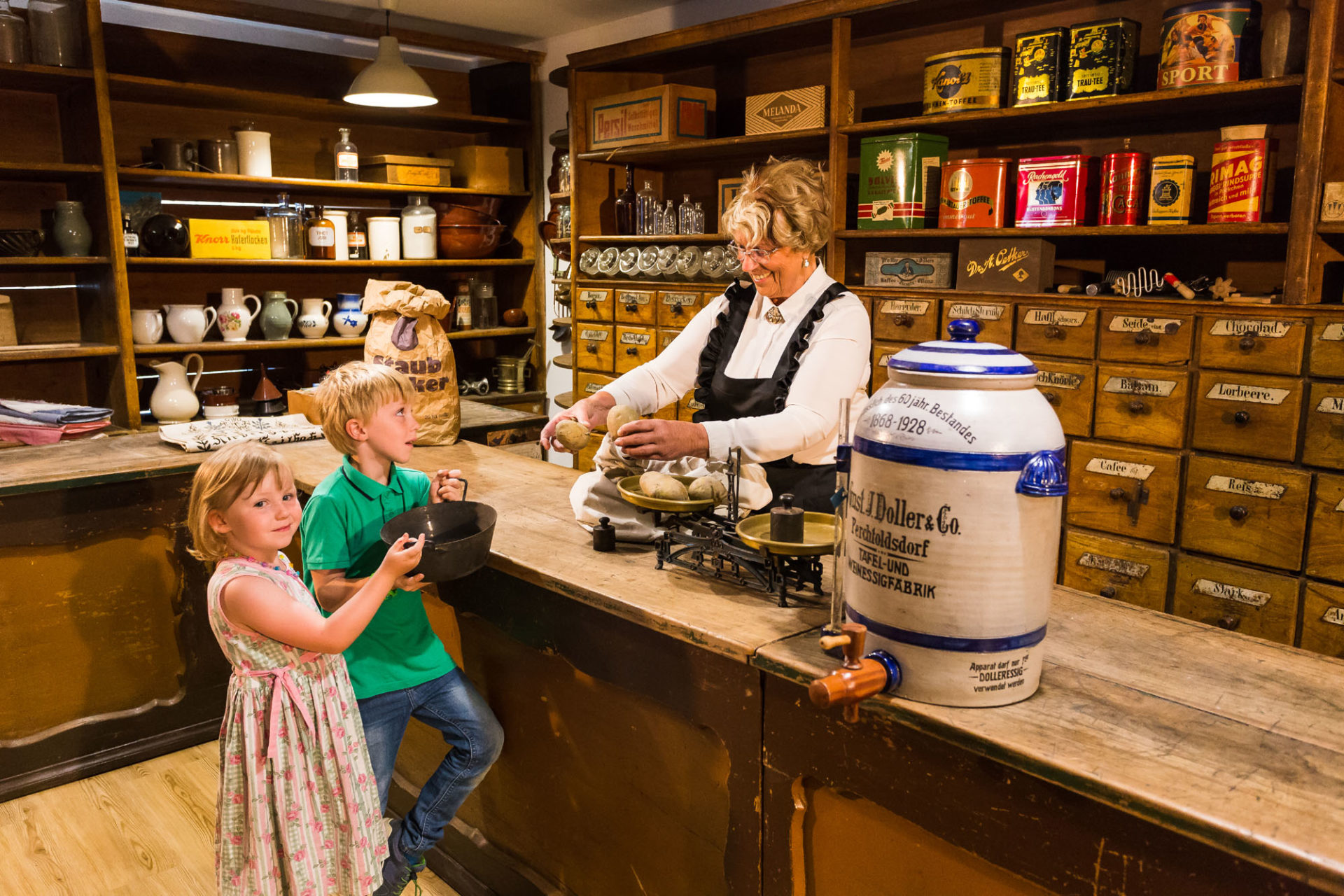 alter Verkaufsladen mit Holzladen im Hintergrund, eine Frau verkauft 2 Kindern Kartoffeln