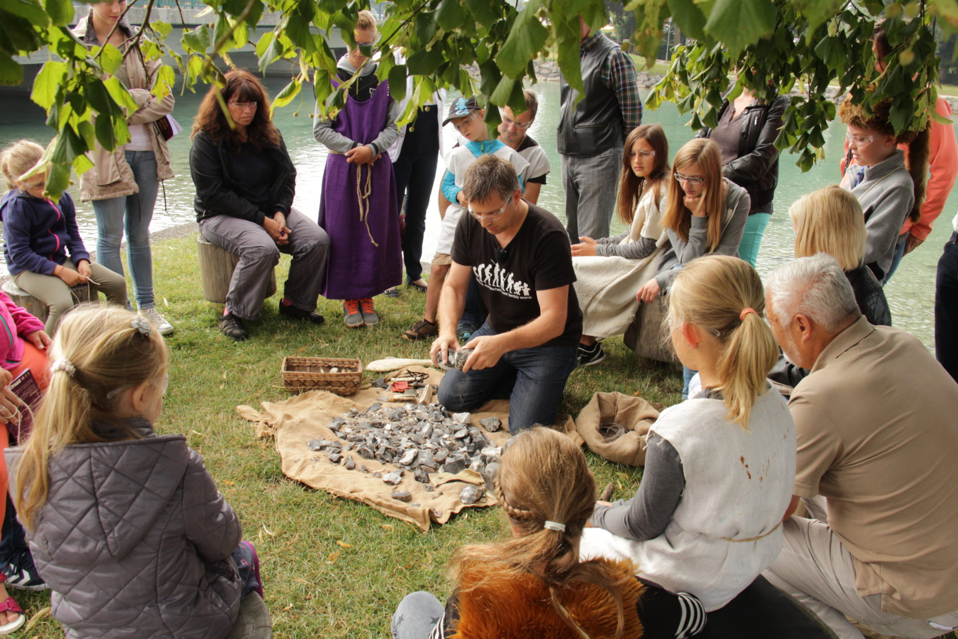 Viele Erwachsene und Kinder sitzen im Kreis um einen Mann der Steine zeigt