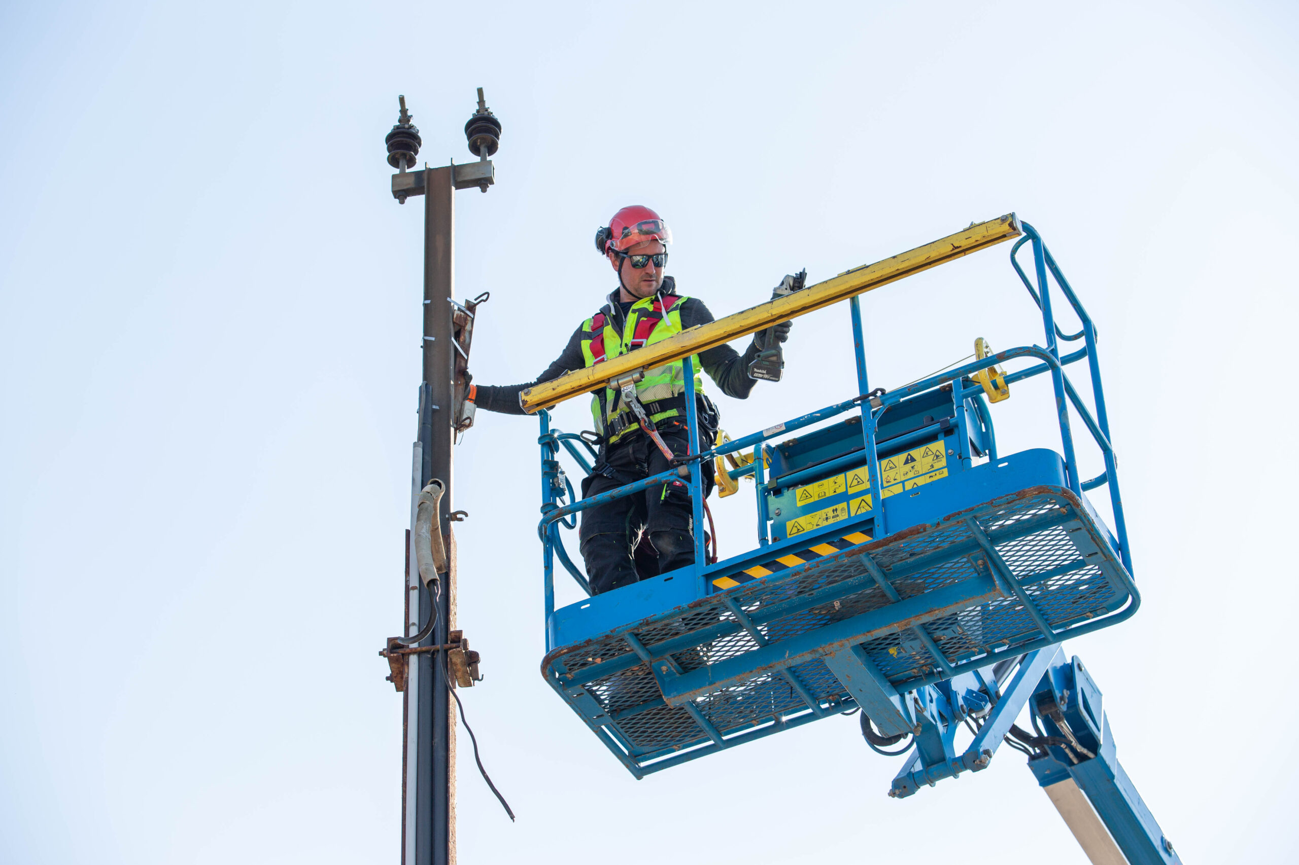 Ein Arbeiter mit gelber Warnweste und Helm steht in einem Kranwagen und arbeitet an einem Fahrleitungsmast
